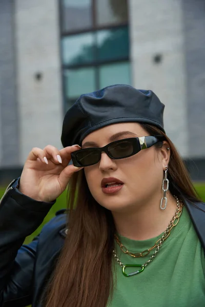 Porträt einer brünetten Frau in Plusgröße, die mit stylischer Sonnenbrille, Lederjacke mit schwarzem Baskenmütze und grüßendem T-Shirt in der Nähe eines verschwommenen modernen Gebäudes auf der städtischen Straße im Freien posiert, körperbetont — Stockfoto