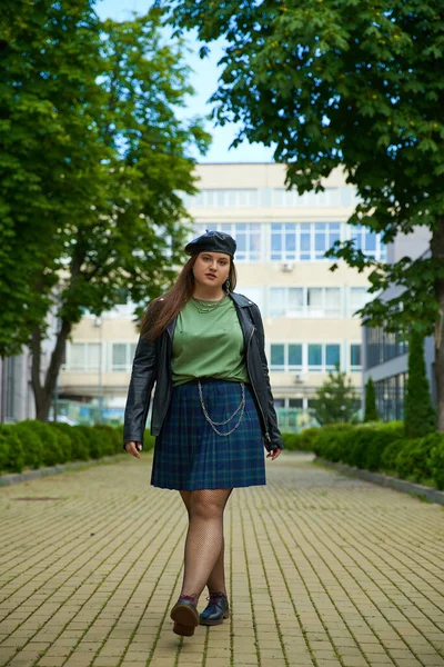 Plus size woman walking in leather jacket, beret, plaid skirt, fishnet tights and black shoes while looking at camera on urban street with building and trees on blurred background, full length — Stock Photo