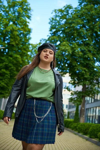 Confident plus size woman walking in leather jacket with black beret, plaid skirt and green t-shirt near blurred modern building on urban street outdoors, body positive, trees on background — Stock Photo