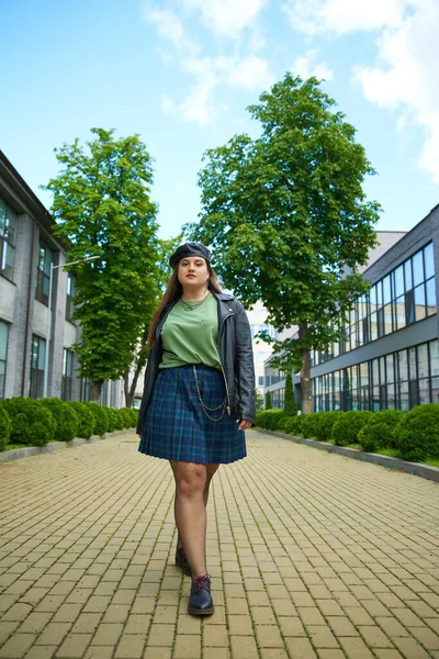 Full length of plus size woman walking in leather jacket, beret, plaid skirt, fishnet tights and black shoes while looking at camera on urban street with buildings and trees on blurred background — Stock Photo