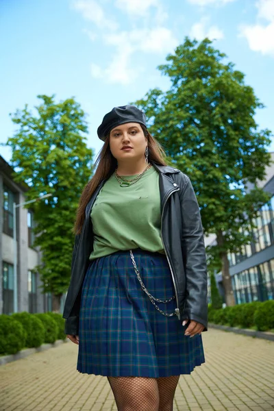 Low angle view of confident woman with plus size body posing in leather jacket with black beret near blurred modern buildings and alley with trees on urban street outdoors, body positive — Stock Photo