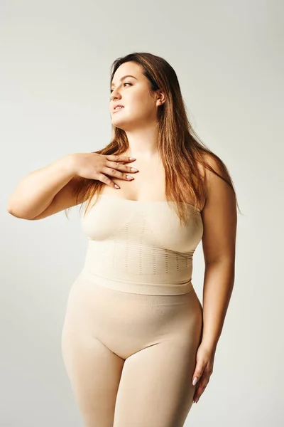 Plus size woman with long hair and bare shoulders looking away while touching chest, posing in beige strapless top and underwear in studio isolated on grey background, body positive, self-love — Stock Photo
