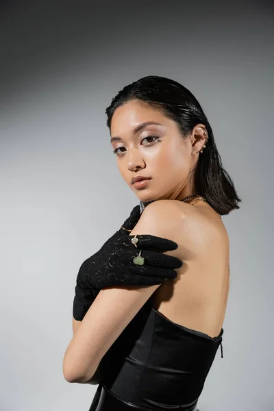 Brunette and asian young woman with short hair posing in black strapless dress and gloves with golden jewelry looking at camera on grey background, wet hairstyle, necklaces, earrings, natural makeup — Stock Photo