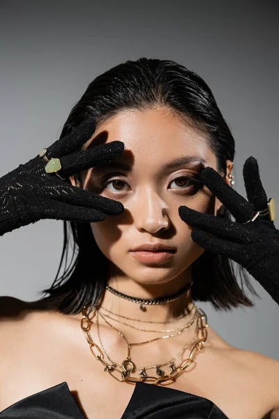 Retrato de morena e asiática jovem mulher com cabelo curto posando em luvas pretas com anéis dourados, olhando para a câmera em fundo cinza, penteado molhado, mãos perto do rosto, maquiagem natural — Fotografia de Stock