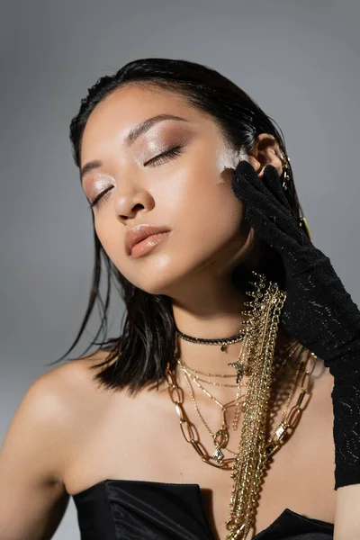 Retrato de morena y mujer joven asiática con pelo corto y ojos cerrados posando en guantes negros y vestido sin tirantes mientras sostiene joyas doradas sobre fondo gris, peinado mojado, maquillaje natural - foto de stock