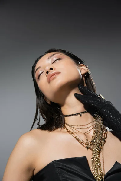 Portrait of alluring and asian young woman with closed eyes and short hair posing in black gloves and strapless dress while holding golden jewelry on grey background, wet hairstyle, natural makeup — Stock Photo