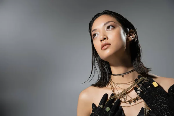 Portrait of mesmerizing asian young woman with short hair posing in black gloves and strapless dress while holding golden jewelry on grey background, wet hairstyle, natural makeup, looking away — Stock Photo