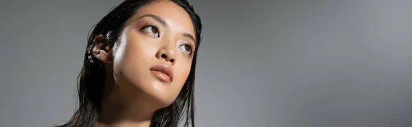 Retrato de fascinante mujer joven asiática con el pelo corto y pendientes de oro posando mientras mira hacia otro lado sobre fondo gris, peinado húmedo, maquillaje natural, mirando hacia otro lado, bandera - foto de stock