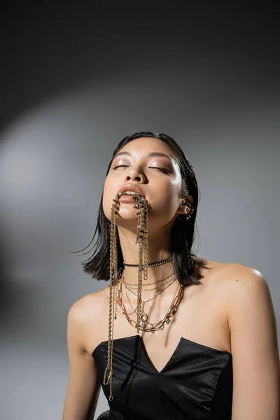 Retrato de mujer joven asiática con pelo corto y ojos cerrados posando en vestido negro sin tirantes mientras sostiene joyas de oro en la boca sobre fondo gris, peinado mojado, maquillaje natural - foto de stock