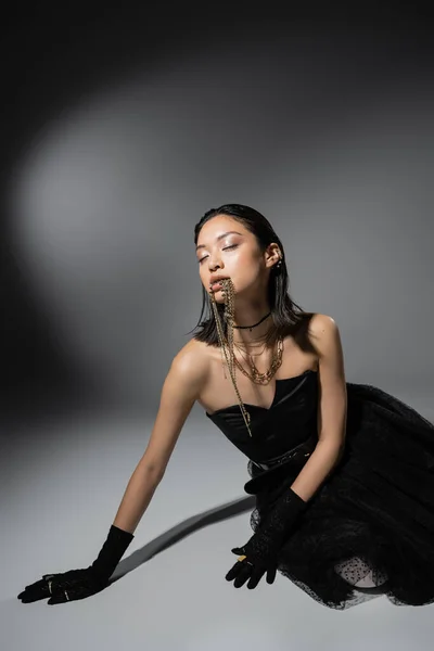 Moda asiática jovem mulher com cabelo curto posando em vestido sem alças preto e luvas enquanto segurando jóias douradas na boca em fundo cinza, maquiagem natural, penteado molhado, olhos fechados — Fotografia de Stock