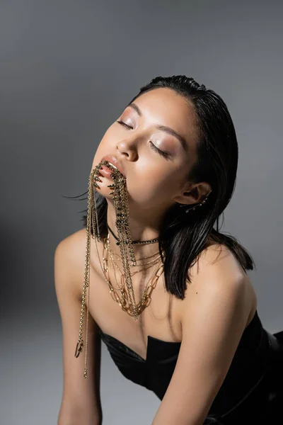 Retrato de moda asiática joven modelo con pelo corto y ojos cerrados posando en negro vestido sin tirantes mientras sostiene joyas de oro en la boca sobre fondo gris, peinado mojado, maquillaje natural - foto de stock