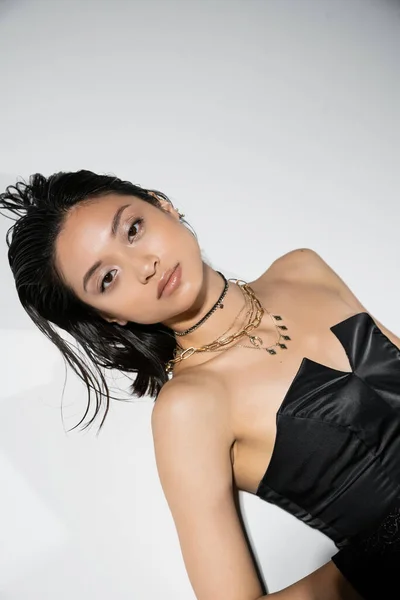 Alluring and asian young woman with short hair lying in black strapless dress while posing in golden jewelry on grey background, wet hairstyle, natural makeup, looking at camera — Stock Photo