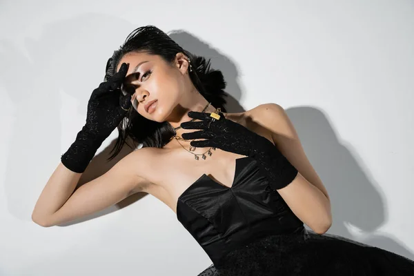 Top view of alluring asian young woman with short hair posing in black gloves and strapless dress, with golden jewelry while lying on grey background, wet hairstyle, natural makeup, looking at camera — Stock Photo