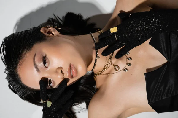 Top view of young asian woman with closed eyes and short brunette hair lying in black gloves and strapless dress while posing in golden jewelry on grey background, wet hairstyle, hand near face — Stock Photo
