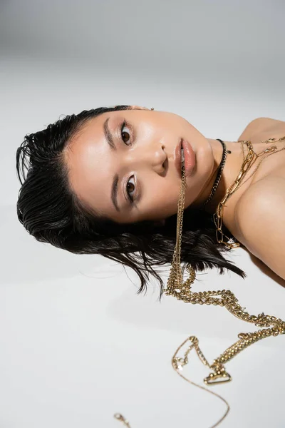 Jovem mulher asiática com cabelo curto morena segurando jóias douradas na boca enquanto olha para a câmera e deitado no fundo cinza, maquiagem cotidiana, penteado molhado, olhos castanhos — Fotografia de Stock
