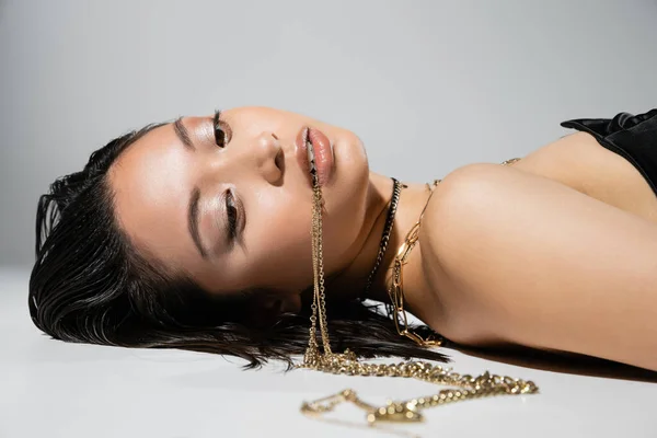 Jovem mulher asiática com cabelo curto morena segurando jóias douradas na boca enquanto posando e deitado no fundo cinza, maquiagem cotidiana, penteado molhado, olhos castanhos — Fotografia de Stock