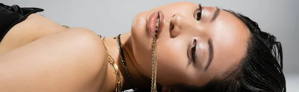Joven mujer asiática con pelo corto morena sosteniendo joyas de oro en la boca mientras posando y acostado sobre fondo gris, maquillaje diario, peinado mojado, ojos marrones, pancarta - foto de stock
