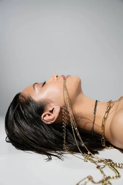 Вид сбоку молодой азиатской женщины с короткими волосами брюнетки, держащей золотые украшения во рту, лежа на сером фоне, повседневный макияж, влажная прическа, закрытые глаза — стоковое фото