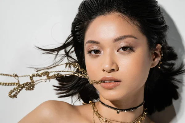 Вид сверху на молодую азиатскую женщину с короткими волосами брюнетки, держащую золотые украшения во рту, глядя в камеру и лежа на сером фоне, повседневный макияж, влажная прическа — стоковое фото