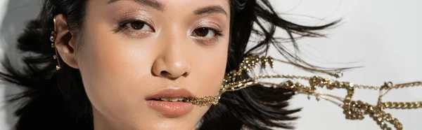 Молодая азиатка с короткими брюнетками, держащая золотые украшения во рту, глядя в камеру и лежа на сером фоне, повседневный макияж, влажная прическа, вид сверху, баннер — стоковое фото