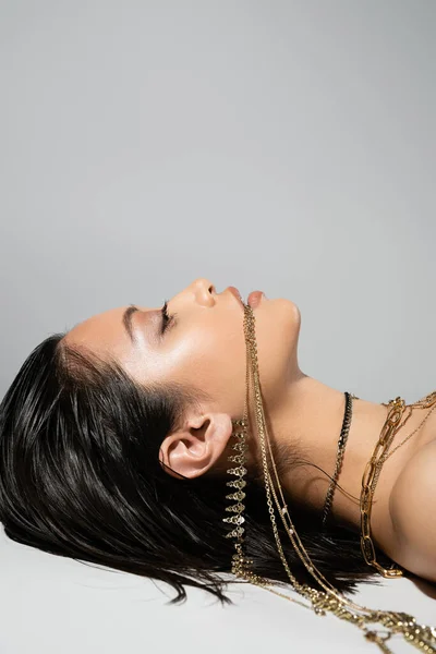 Jovem mulher asiática com cabelo curto morena segurando jóias douradas na boca enquanto posando em fundo cinza, maquiagem cotidiana, penteado molhado, vista lateral, olhando para cima — Fotografia de Stock