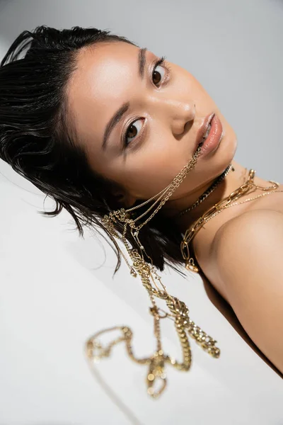 Asian model with short brunette hair holding golden jewelry in mouth while looking at camera and lying on grey background, everyday makeup, wet hairstyle, young woman — Stock Photo