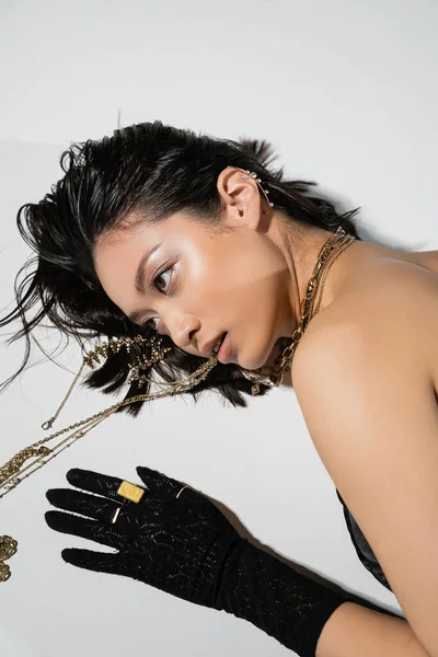 Overhead view of asian model with short brunette hair holding golden jewelry in mouth while looking away and lying on grey background, everyday makeup, wet hairstyle, young woman, black gloves — Stock Photo