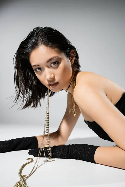 Asian model with short brunette hair holding golden jewelry in mouth while looking at camera and posing in strapless dress on grey background, everyday makeup, wet hairstyle, young woman — Stock Photo