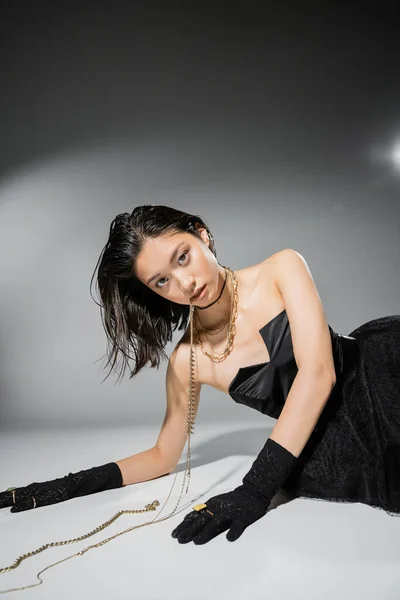 Asian model with short brunette hair holding golden jewelry in mouth while looking at camera and posing in black strapless dress on grey background, everyday makeup, wet hairstyle, young woman — Stock Photo
