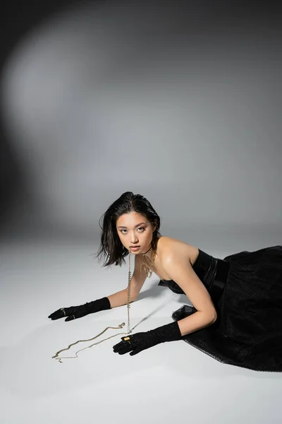 Modelo asiático de moda con pelo corto morena celebración de joyas de oro en la boca mientras mira a la cámara y posando en vestido sin tirantes sobre fondo gris, maquillaje diario, peinado húmedo, mujer joven - foto de stock