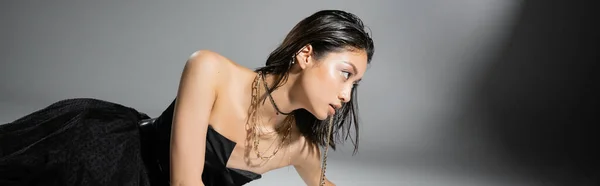 Asian model with short brunette hair holding golden jewelry in mouth while looking away and posing in strapless dress on grey background, everyday makeup, wet hairstyle, young woman, banner — Stock Photo