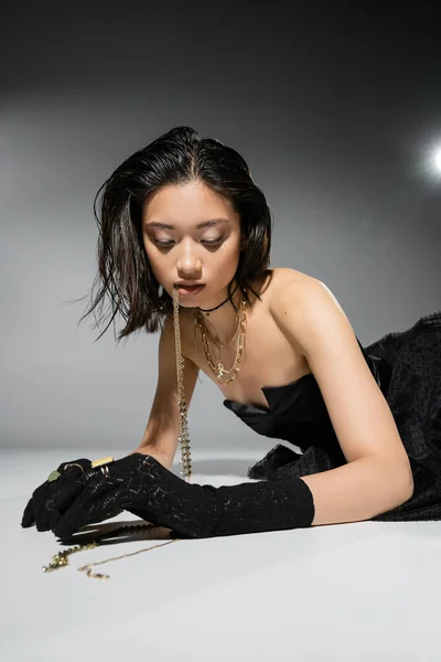 Captivating asian model with short brunette hair holding golden jewelry in mouth while looking down and posing in strapless dress on grey background, everyday makeup, wet hairstyle, young woman — Stock Photo