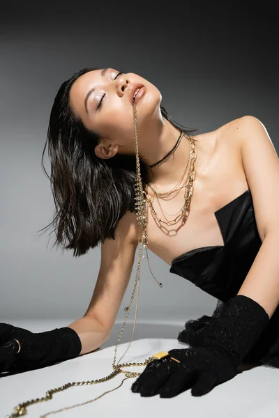 Encantador modelo asiático com cabelo curto morena segurando jóias douradas na boca enquanto posando em vestido sem alças no fundo cinza, maquiagem cotidiana, penteado molhado, jovem mulher, olhos fechados — Fotografia de Stock