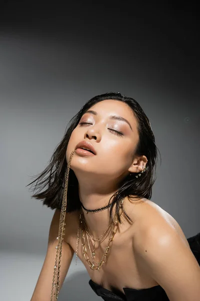 Stylish asian model with short brunette hair holding golden jewelry in mouth, posing in strapless dress on grey background, everyday makeup, wet hairstyle, young woman, closed eyes, portrait — Stock Photo