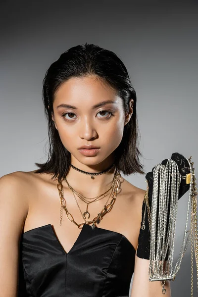 Portrait of brunette and asian young woman with short hair holding golden and silver jewelry while wearing glove and standing in strapless dress on grey background, wet hairstyle, natural makeup — Stock Photo