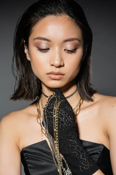 Portrait of alluring asian young woman with short hair holding golden and silver jewelry while wearing glove and standing in strapless dress grey background, wet hairstyle, natural makeup — Stock Photo