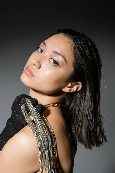 Портрет соблазнительной азиатской молодой женщины с короткими волосами, позирующей в черной перчатке и держащей золотые и серебряные украшения на плече, стоя на сером фоне, влажная прическа, натуральный макияж — стоковое фото