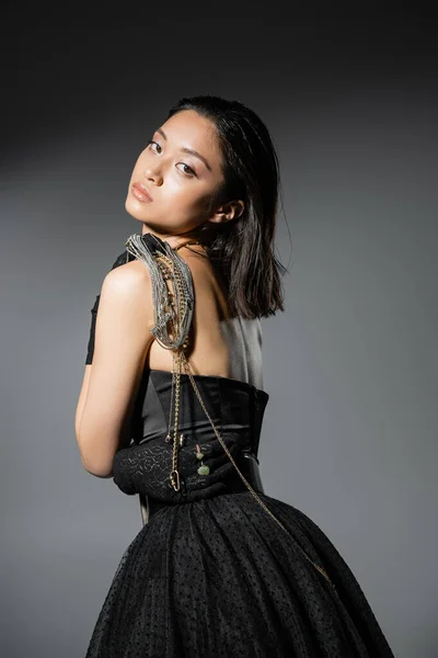 Portrait of brunette and asian young woman with short hair posing with golden and silver jewelry on shoulder while standing in black strapless dress on grey background, wet hairstyle, natural makeup — Stock Photo