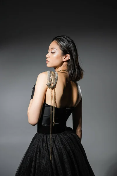 Side view of brunette and asian young woman with short hair posing with golden and silver jewelry on shoulder while standing in strapless dress on grey background, wet hairstyle, natural makeup — Stock Photo