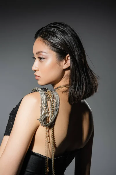 Portrait of brunette and asian young woman with short hair posing with golden and silver jewelry on shoulder while standing in strapless dress on grey background, wet hairstyle, natural makeup, side view — Stock Photo