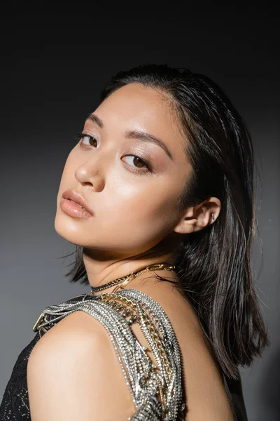 Portrait of brunette and asian young woman with short hair posing with golden and silver jewelry on shoulder and looking at camera on grey background, wet hairstyle, natural makeup — Stock Photo