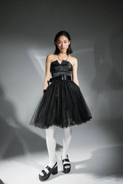 Full length of stylish asian young woman with short hair posing in black strapless dress, white tights and shoes while looking at camera on grey background, wet hairstyle, golden necklaces — Stock Photo
