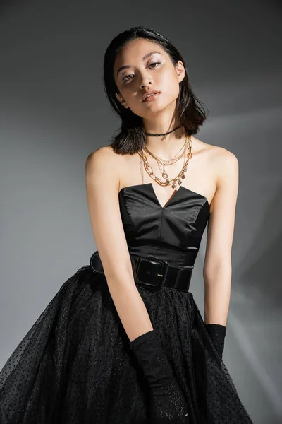 Портрет стильної азіатської молодої жінки з коротким волоссям, що позує в чорній сукні з поясом і рукавичками, дивлячись на камеру на сірому фоні, мокру зачіску, золоті намиста — стокове фото