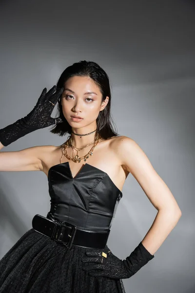 Mesmerizing young asian woman with short hair posing with hand on hip in black strapless dress with belt and gloves while looking at camera on grey background, wet hairstyle, golden necklaces — Stock Photo