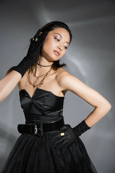 Mujer asiática joven glamorosa con pelo corto posando con la mano en la cadera en vestido negro sin tirantes con cinturón y guantes mientras mira hacia otro lado sobre fondo gris, peinado mojado, collares dorados - foto de stock