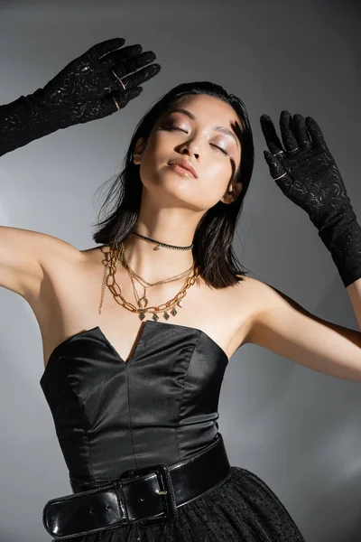 Стильная азиатская молодая женщина с короткими волосами позирует с поднятыми руками в черном платье без бретелек с поясом и перчатками на сером фоне, влажная прическа, золотые ожерелья, закрытые глаза — стоковое фото