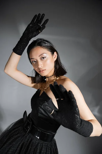 Mujer joven asiática glamorosa con pelo corto posando con las manos levantadas en vestido negro sin tirantes con cinturón y guantes sobre fondo gris, peinado mojado, collares dorados, mirando a la cámara - foto de stock