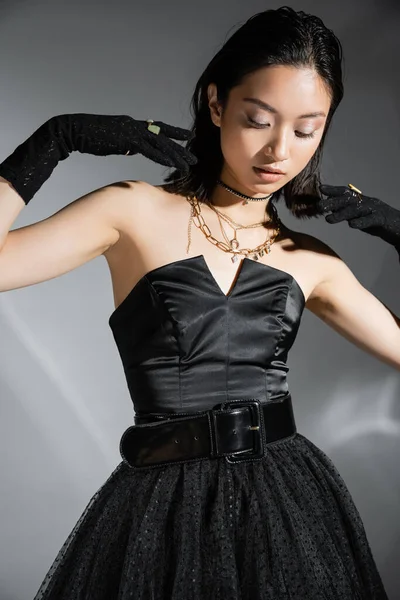 Glamorosa mujer joven asiática con pelo corto posando en vestido negro sin tirantes con cinturón y guantes mientras mira hacia abajo sobre fondo gris, peinado mojado, collares dorados - foto de stock