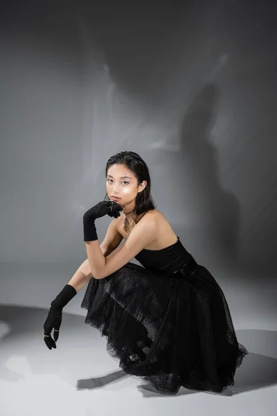 Full length of stylish asian young woman with short hair sitting in black strapless dress with tulle skirt and gloves while looking at camera on grey background, wet hairstyle, golden necklaces — Stock Photo