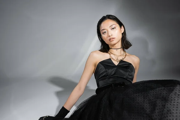 Грациозная азиатская женщина с короткими волосами, сидящая в черном платье без бретелек с юбкой тюля, пояс при взгляде на камеру на сером фоне, влажная прическа, золотые ожерелья, завораживающая красота — стоковое фото